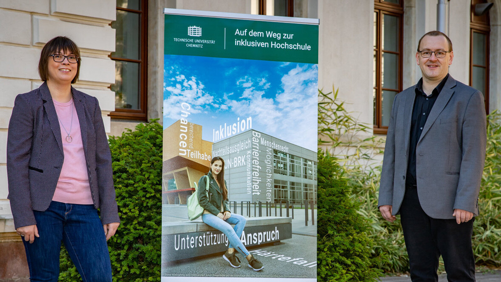 EIne Frau und ein Mann in Blazer und Sakko stehen neben einem Schild. Darauf steht Inkluaisve Hochschule.