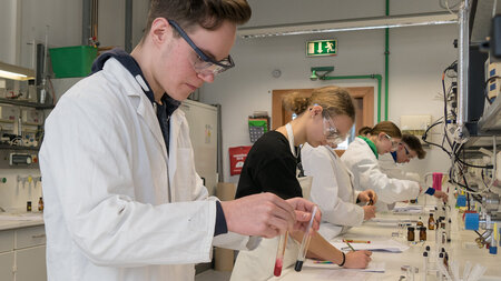 Schülerinnen und Schüler in weißen Kitteln hantieren mit Reagenzien an Labortischen.