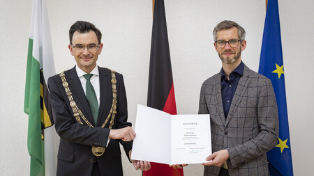 A man in a suit and a big chain and a man in a jacket with glasses are holding a certificate together.