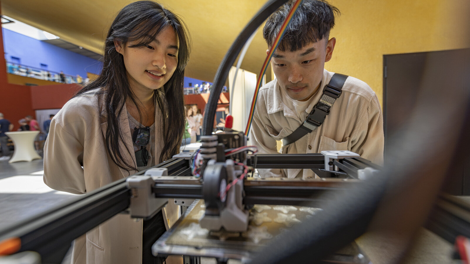 Eine junde Frau und ein junger Mann blicken in einen 3D-Drucker.