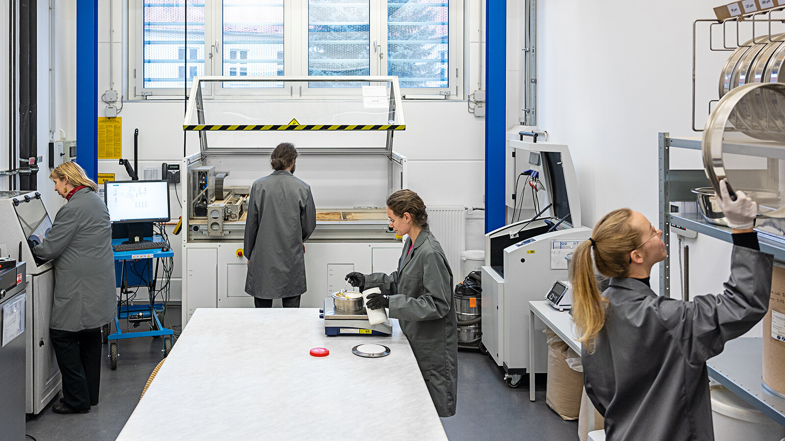 Vier Personen in grauen Kitteln arbeiten an verschiedenen Stationen in einem Labor. 