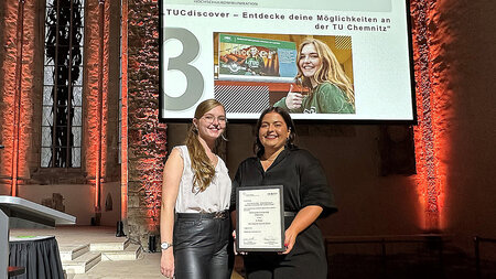 Two young women are standing next to each other. One is holding a certificate in her hand. Behind them on a screen the number 3 is visible.