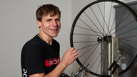 Ein lächelnder Mann steht neben einem an einer Werkbank eingespannten Vorderrad eines Fahrrades.