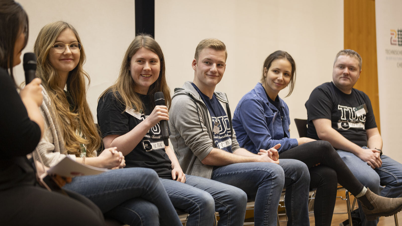 Fünf Studierende sitzen nebeneinander auf Stühlen, eine Person hält ein Mikrofon in der Hand.