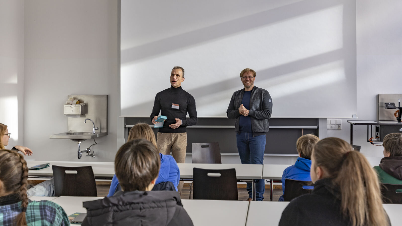 Zwei Männer stehen vor einer Tafel und sprechen mit Personen im Seminarraum.