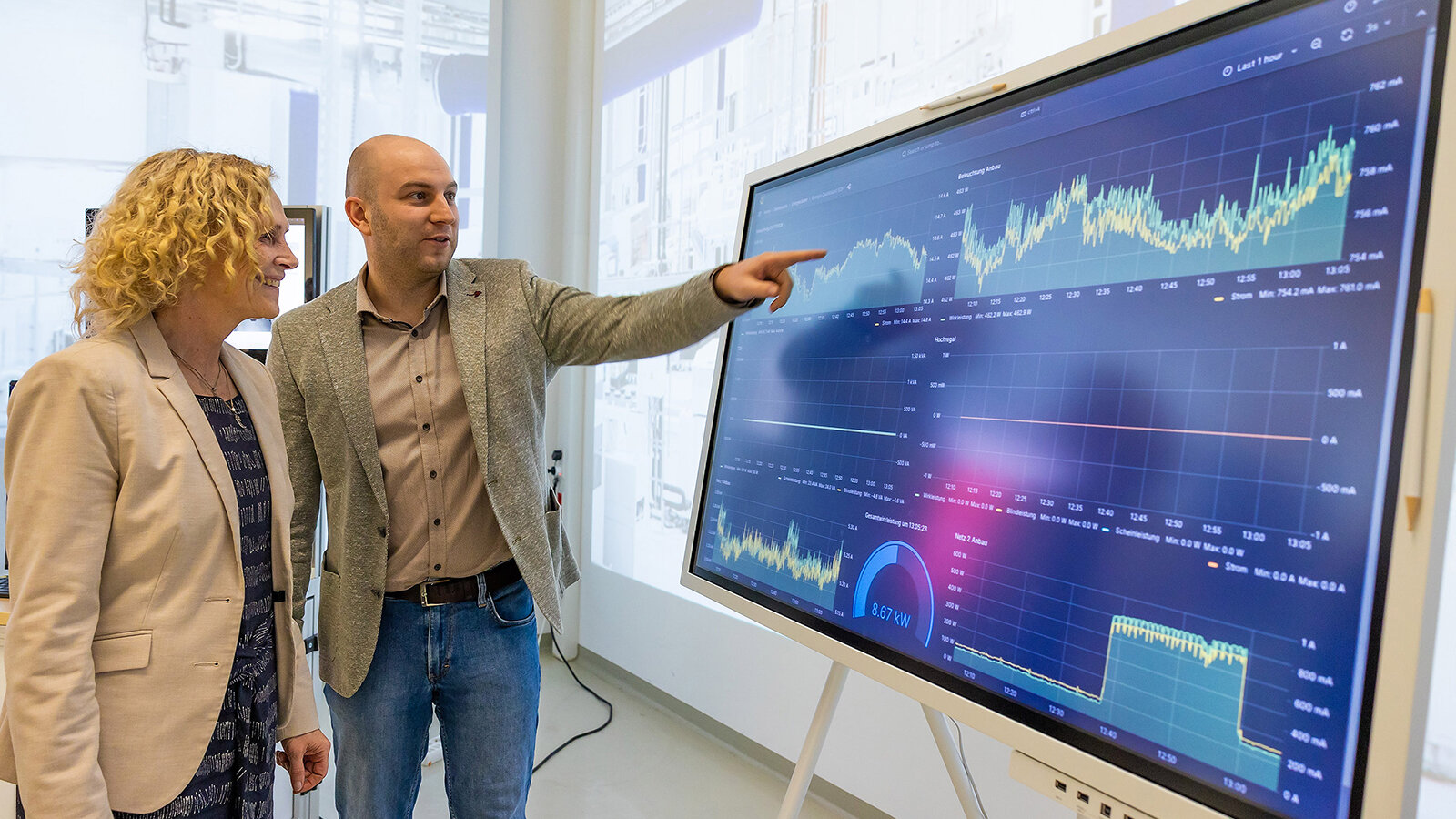 Ein Mann zeigt einer Frau auf einem großen Monitor Diagramme.