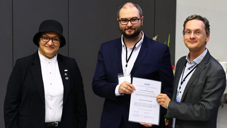 Eine Frau und zwei Männer stehen nebeneinander. Ein Mann übergibt eine Urkunde.