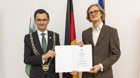 Zwei Männer stehen nebeneinander und halten eine Urkunde in den Händen.