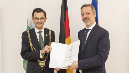 Zwei Männer stehen nebeneinander und halten eine Urkunde in den Händen.
