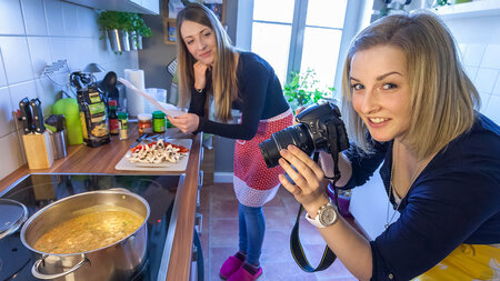 Zwei junge Frauen agieren in einer Küche, eine fotografiert das Essen auf dem Herd.