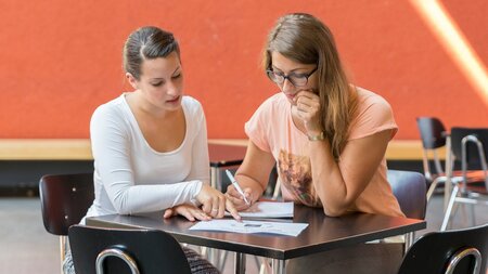 Zwei Frauen sitzen an einem Tisch und schauen auf Faltblätter.