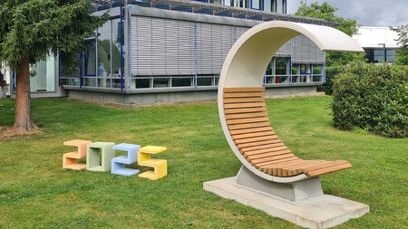 Ein C-förmiges Sitzmöbel steht auf einem Rasen vor einem Gebäude, daneben kleinere Hocker.