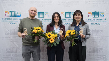Ein Mann und zwei Frauen stehen nebeneinander. IN den Hnden halten sie einen kleinen hlzernen Pokal und einen Blumenstrau.