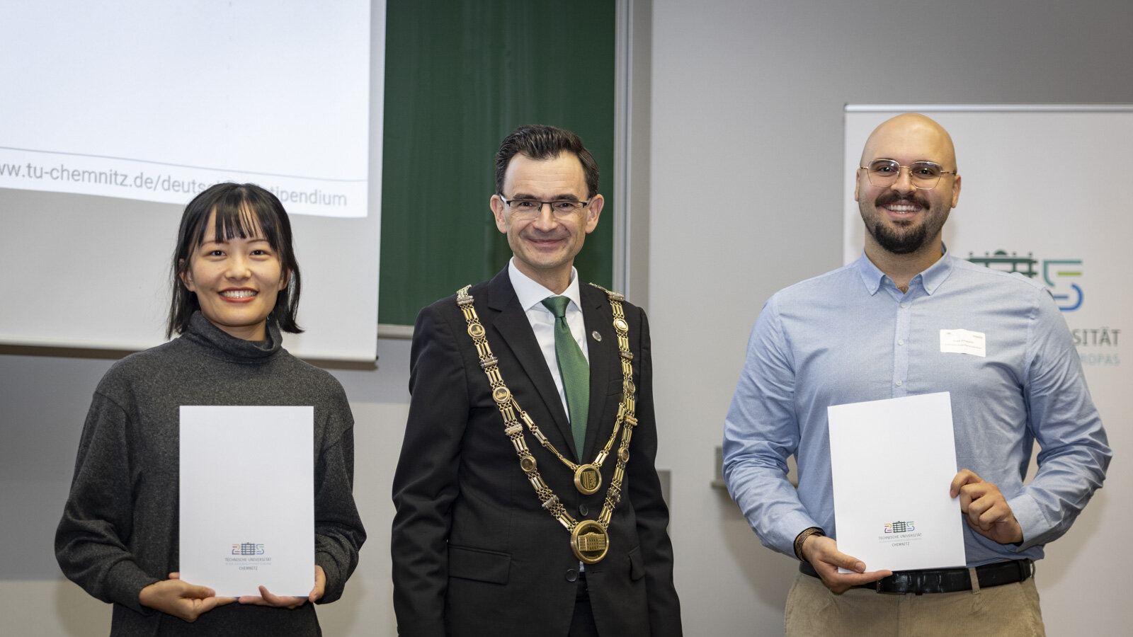 Ein Mann mit Amtskette steht zwischen einer Frau und einem Mann.