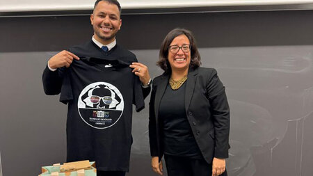 A man holding up a T-shirt stands next to a woman in front of a blackboard. 