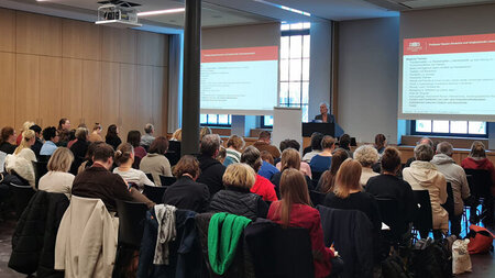 Mehrere Personen sitzen in einem Raum und blicken zu einer Person, die einen Vortrag hält.