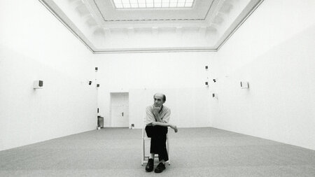 Ein Mann sitzt in einem großen hellen Raum mit kleinen Lautsprechern an den Wänden auf einen Stuhl