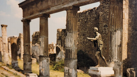 Eine Skulptur steht vor Ruinen eines Hauses mit Säulen. 
