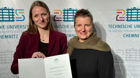 Zwei Frauen stehen nebeneinander vor einer Messewand, eine Frau hält eine Urkunde in den Händen. 