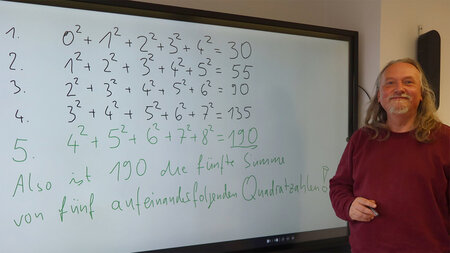 Ein Mann steht neben einem großen Monitor, auf dem Formeln zu sehen sind.