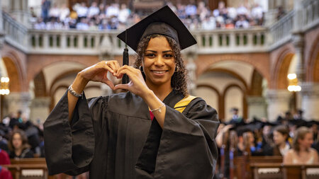 Eine Frau im Talar steht in einer Kirche und symbolisiert mit ihren Händen ein Herz.