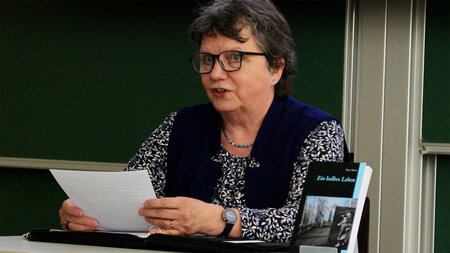 Eine Frau sitzt an einem Tisch und liest aus einem Buch vor.