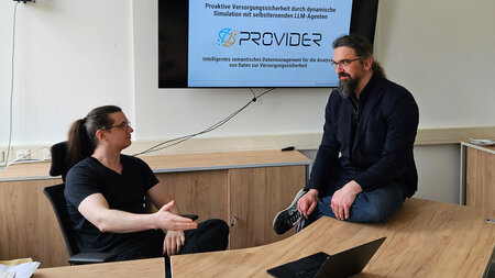 Zwei Männer sitzen an beziehungsweise auf einem Tisch und sprechen miteinander.