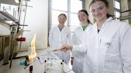 BildbeschDrei Mädchen stehen in einer Reihe vor einem Experiment