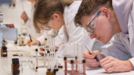 Schülerinnen und Schüler in weißen Kitteln experimentieren an einem Labortisch mit Reagenzien. 