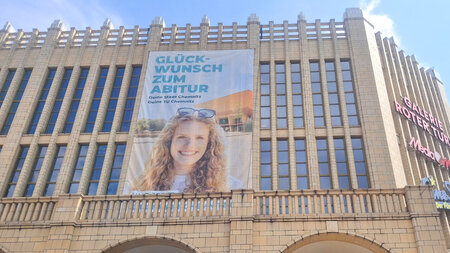 An einem Gebäude ist ein riesiges Banner befestigt, das eine lächelnde Frau vor einem Haus zeigt und Werbebotschaften enthält.