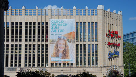 An einem Gebäude ist ein riesiges Banner befestigt, das eine lächelnde Frau vor einem Haus zeigt und Werbebotschaften enthält.