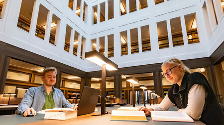 Ein Mann und Frau sitzen in einem Lesesall an einem Tisch und lesen in Büchern.
