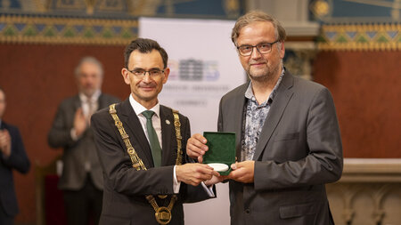 Ein Mann mit Amtskette überreich einem anderen Mann eine Medaille.
