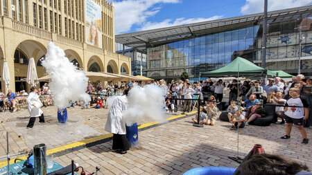 Mehrere Personen beobachetn Chemieexperimente auf einem Marktplatz.