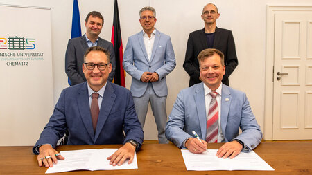 Zwei Männer sitzen an einem Tisch und unterzeichnen Dokumente. Drei Männer stehen dahinter. 