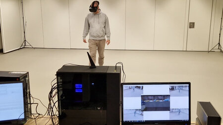 Ein Mann mit VR-Brille auf dem Kopf steht in einem hellen Raum hinter Tischen, auf denen Computer stehen.