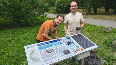 Zwei Männer monieren eine Box an eine Informationstafel, die an einem Wanderweg steht.