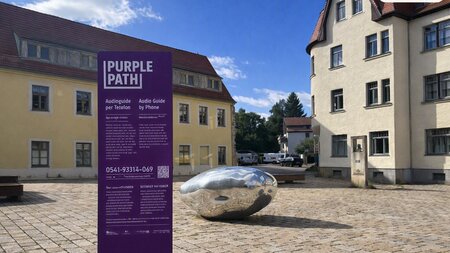 Eine beschriftete Stele steht neben einem silbernen Metallklumpen auf einm Platz vor einem Gebäude.