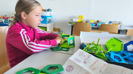 Ein Mädchen sitzt an einem Tisch und bastelt mit bunten Plastikteilen. 