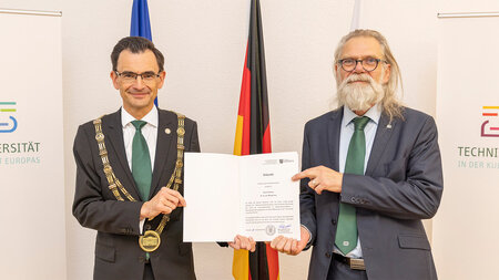 Zwei Männer im Anzug stehen nebeneinander und halten eine Urkunde in den Händen.