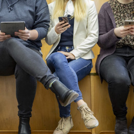 Three persons sitting in a lecture room holding smart phones and tablets in their hands.
