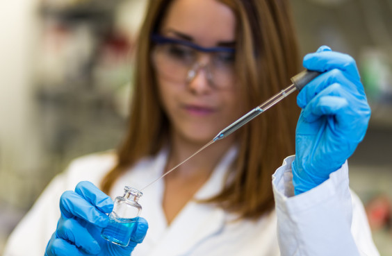 Wissenschaftlerin in einem Laborkittel entnimmt mit einer Pipette Flüssigkeit aus einem kleinen Glas