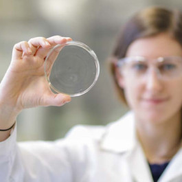 Chemikerin in Laborkittel und Schutzbrille, die eine Petrischale hochhält