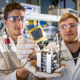 Two chemists in lab coats and safety goggles holding up a fuel cell